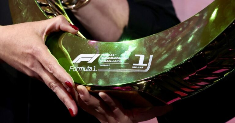 Mãos segurando o troféu do primeiro lugar do Grande Prêmio de São Paulo de Fórmula 1 de 2024, patrocinado pela Lenovo. O troféu possui um formato circular e metálico, com acabamento em tom dourado e reflexos multicoloridos. No centro, está o logotipo da Fórmula 1 ao lado da inscrição "Lenovo Grande Prêmio de São Paulo 2024 – São Paulo – First Place".