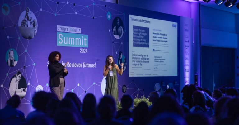 Palestrante no palco do Programaria Summit 2024, ao lado de uma intérprete de libras, aborda o tema da utilização de inteligência artificial para a criação de conteúdo indevido. No telão ao fundo, são exibidas manchetes relacionadas a problemas de moderação de conteúdo de abuso infantil e uso de IA. O cenário é composto por luzes em tons de azul e púrpura, com público atento acompanhando a apresentação.