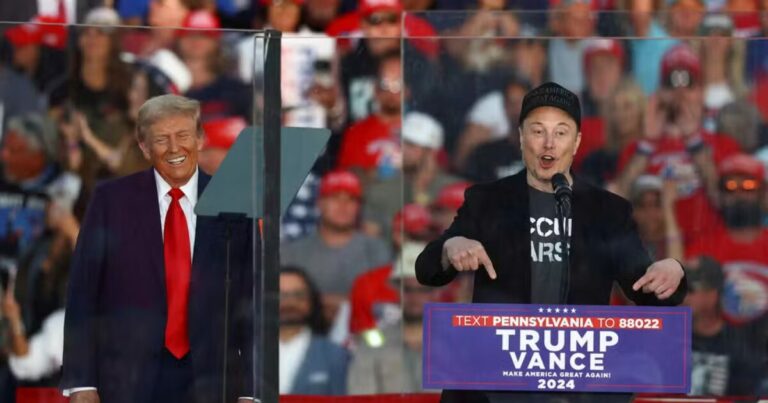 Elon Musk e Donald Trump em um comício de apoio ao candidato republicano J.D. Vance na Pensilvânia, EUA. Trump sorri ao fundo enquanto Musk, com boné e camiseta, faz gestos com as mãos ao falar ao microfone, com uma multidão ao fundo.