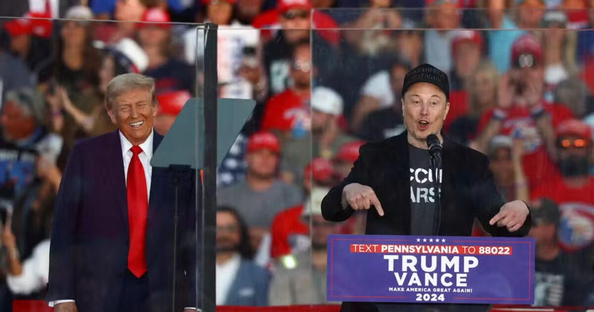 Elon Musk e Donald Trump em um comício de apoio ao candidato republicano J.D. Vance na Pensilvânia, EUA. Trump sorri ao fundo enquanto Musk, com boné e camiseta, faz gestos com as mãos ao falar ao microfone, com uma multidão ao fundo.