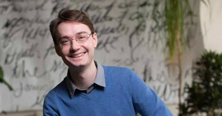 François Chollet usando óculos de aro fino e uma camisa azul, está sorrindo para a câmera. O fundo é artístico, com uma parede coberta por textos manuscritos e plantas, dando um tom informal e criativo à imagem. Ele parece relaxado e amigável, com um visual que sugere um ambiente profissional descontraído.