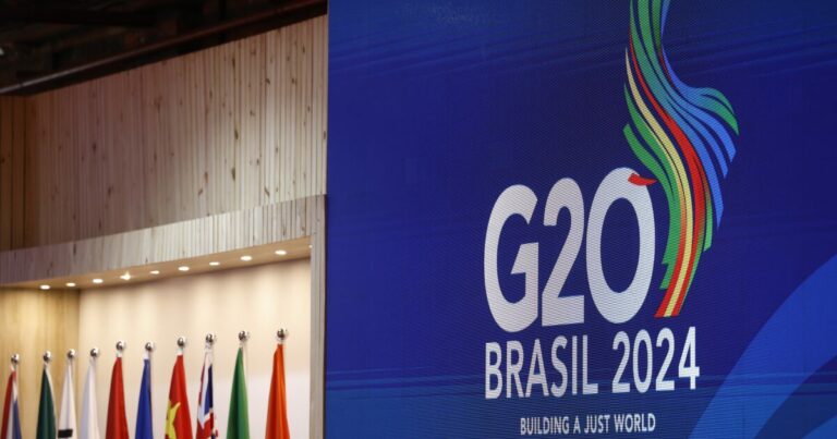 Imagem do evento G20 Brasil 2024, com um painel azul exibindo o logo oficial do evento, composto pelo texto 'G20 Brasil 2024' e o slogan 'Building a Just World'. Ao lado, há bandeiras dos países membros alinhadas em um ambiente com parede de madeira.