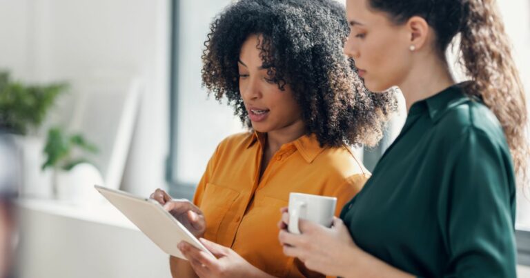 A imagem mostra duas mulheres em um ambiente de trabalho. A mulher à esquerda, que tem cabelo cacheado volumoso e usa uma camisa amarela, está segurando um tablet e explicando algo à outra mulher. A mulher à direita, que tem cabelo preso em um rabo de cavalo e veste uma camisa verde-escura, está segurando uma caneca branca enquanto observa atentamente a tela do tablet. O cenário ao fundo é iluminado, com tons claros, sugerindo um ambiente profissional ou de escritório.