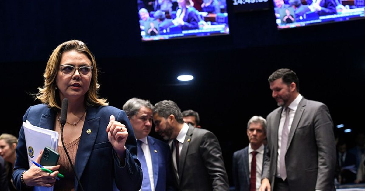 Uma mulher, usando óculos e um blazer azul, está falando em um microfone em um ambiente formal. Ela segura documentos e um celular na mão. Atrás dela, há outras pessoas, incluindo homens em trajes formais, conversando entre si. No fundo, vê-se telas exibindo imagens do mesmo local, sugerindo que o evento ocorre em um plenário ou sessão legislativa, lei