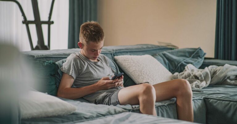 Adolescente sentado em um sofá, usando um smartphone para acessar suas redes sociais, em um ambiente doméstico com almofadas e uma manta ao fundo. (Google, Meta, Character.AI)