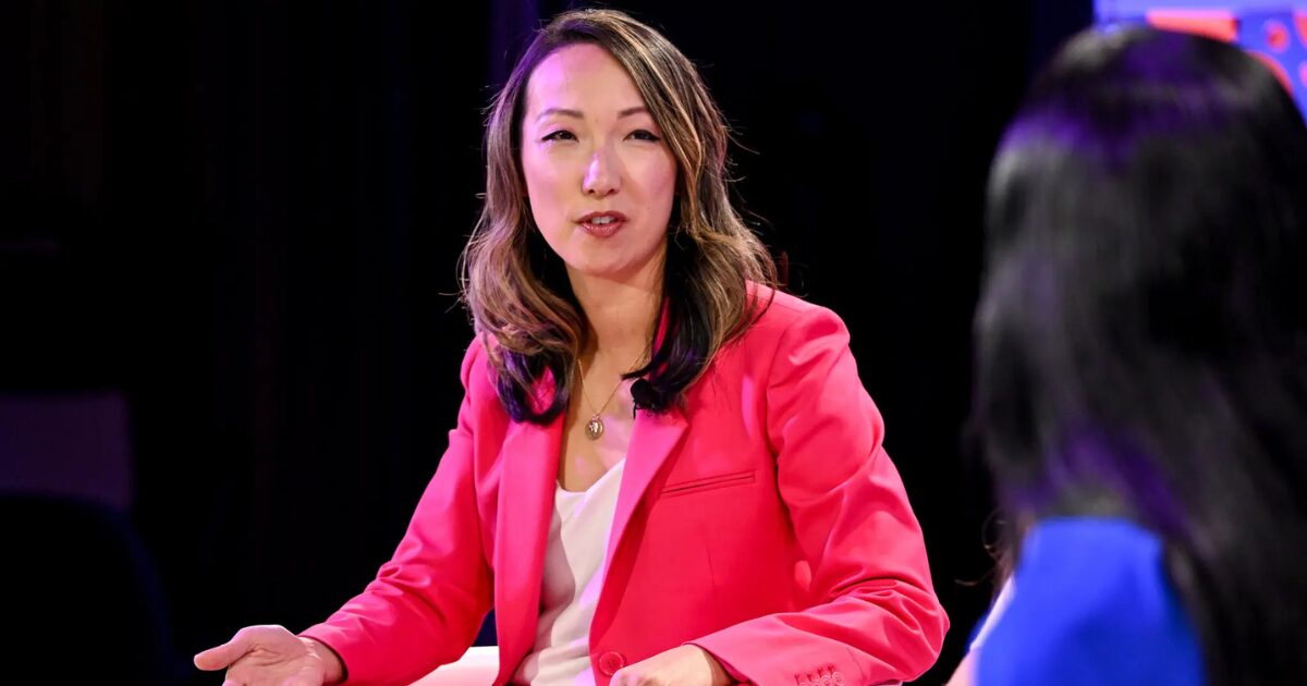 Imagem de uma mulher vestindo um blazer rosa, participando de uma conversa em um ambiente fechado. Ela tem cabelos longos e ondulados, e está falando enquanto gesticula com a mão esquerda. A iluminação do ambiente destaca o rosto dela, e ao fundo há uma outra pessoa, parcialmente desfocada, com o cabelo escuro. A expressão facial da mulher indica um tom de seriedade e atenção. (Meta)