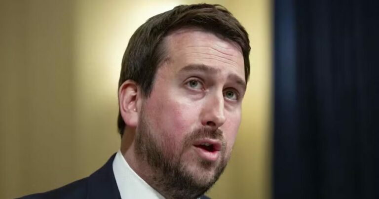 A imagem mostra Nick Pickles com cabelos curtos e barba curta, usando um terno escuro e gravata, falando enquanto olha ligeiramente para o lado. O fundo é neutro, com iluminação suave, destacando o rosto da pessoa na imagem.