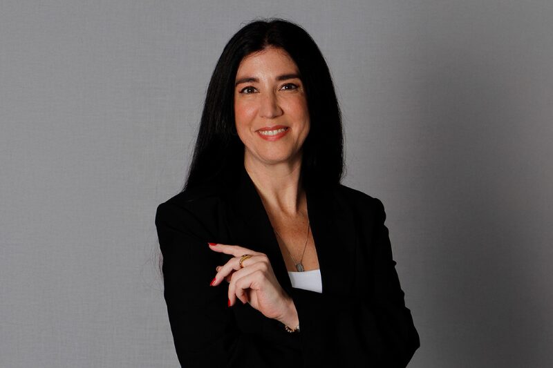 Retrato profissional de uma mulher de cabelos escuros e longos, vestindo um blazer preto sobre uma blusa branca. Ela está de frente para a câmera, com uma expressão amigável e confiante, com os braços cruzados levemente em frente ao corpo. O fundo é neutro, destacando o foco na pessoa retratada, Pierella Paolada da Tivit