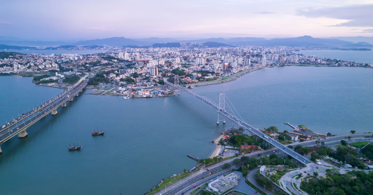 A imagem mostra uma vista aérea da cidade de Florianópolis, com destaque para a Ponte Hercílio Luz, que conecta a ilha de Florianópolis ao continente. A ponte aparece no centro da imagem, cruzando o mar. Ao fundo, pode-se ver a área urbana da cidade com vários prédios altos, além de montanhas ao longe. O cenário é composto por águas calmas, com algumas embarcações visíveis, e a iluminação suave do entardecer cria uma atmosfera tranquila e cênica