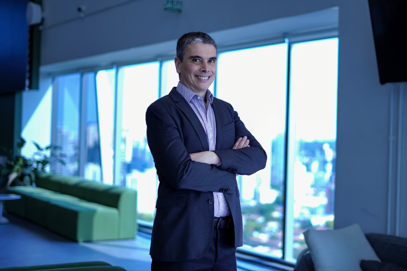 Foto de Rodrigo Couto, Vice-Presidente de Finanças do PicPay, em um ambiente corporativo moderno. Ele está de braços cruzados e veste um terno escuro com camisa clara, sorrindo para a câmera. Ao fundo, grandes janelas oferecem uma vista ampla da cidade, complementando a atmosfera profissional e elegante