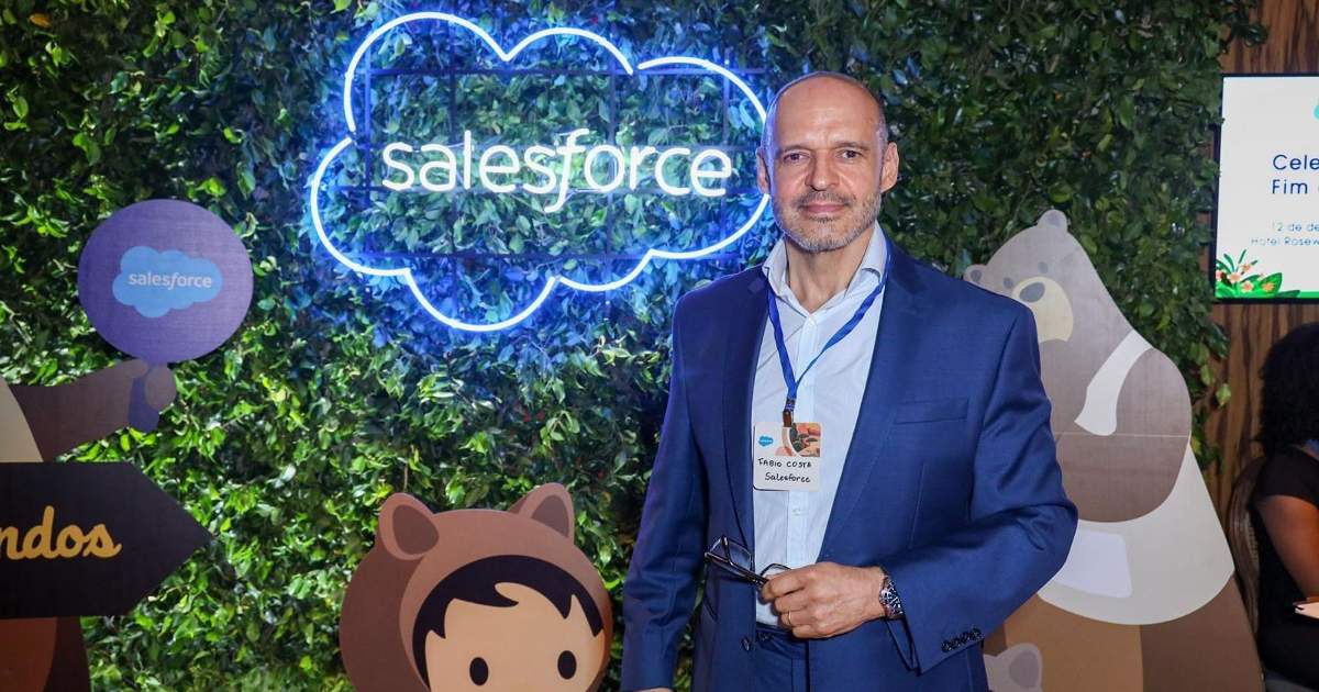 Foto de Fabio Costa, ex-general manager da Salesforce no Brasil, em um evento da empresa. Ele está vestido com um blazer azul e camisa clara, usando um crachá de identificação. Ao fundo, há uma parede de folhagens com o logotipo iluminado da Salesforce, além de figuras de mascotes característicos da empresa, criando um ambiente descontraído e temático