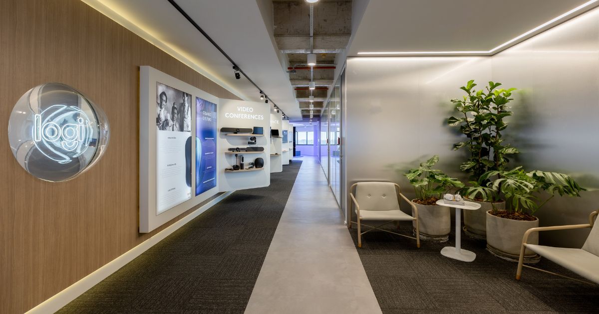 A entrada do showroom da Logitech em São Paulo transmite um ambiente moderno e sofisticado. À esquerda, o logo iluminado da "Logi" se destaca em uma parede de madeira clara, trazendo uma atmosfera acolhedora. Mais adiante, há uma seção de produtos voltados para videoconferência, exibidos em prateleiras suspensas e acompanhados por painéis com informações detalhadas, reforçando o compromisso da marca com tecnologia e inovação.

O corredor espaçoso segue em direção a áreas mais abertas, com iluminação indireta e design clean, combinando piso em concreto polido e carpete escuro. À direita, um espaço de espera confortável foi decorado com poltronas de design minimalista e vasos com plantas verdes, criando uma atmosfera de aconchego e frescor. Esse layout reflete a identidade da Logitech, unindo tecnologia e bem-estar em um ambiente funcional e esteticamente agradável.