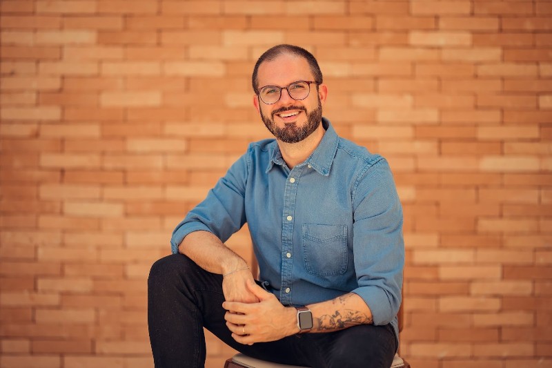 Foto de Luis Felipe Fernandes, CEO da Kokedama AI Studio, sentado casualmente em frente a uma parede de tijolos. Ele está sorrindo, veste uma camisa jeans azul e calça preta, usa óculos redondos e tem uma barba bem cuidada. Um relógio inteligente no pulso e uma tatuagem no braço completam seu visual moderno e descontraído, transmitindo confiança e simpatia