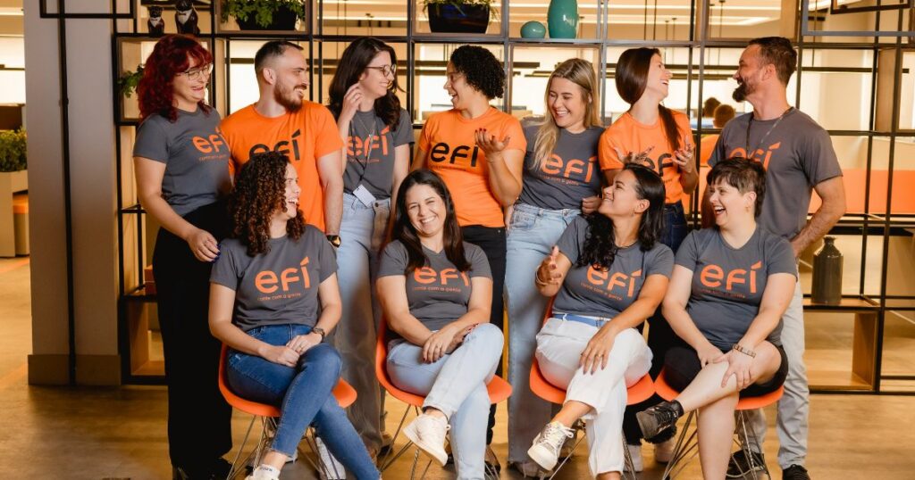 Nesta foto, vemos o time da Éfi em um momento descontraído, demonstrando claramente a camaradagem e o espírito colaborativo da equipe. Todos estão vestidos com camisetas da marca Éfi, nas cores laranja e cinza, criando um visual harmonioso e coeso. O grupo está sorrindo, trocando olhares e gesticulando, indicando uma atmosfera alegre e de integração. A imagem transmite um ambiente de trabalho acolhedor e descontraído, onde todos parecem à vontade e conectados uns com os outros. A disposição informal e os sorrisos refletem um time engajado e unido, com forte senso de equipe.