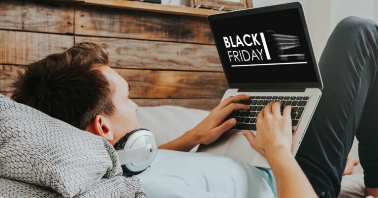A imagem mostra uma pessoa relaxando em uma cama, usando um laptop e com fones de ouvido ao redor do pescoço. No laptop, é possível ver a tela com o texto "BLACK FRIDAY", indicando que a pessoa está navegando em ofertas da Black Friday. O ambiente ao fundo possui uma parede de madeira, criando uma atmosfera aconchegante. A pessoa está usando uma camiseta clara e parece focada na tela do computador enquanto aproveita as promoções