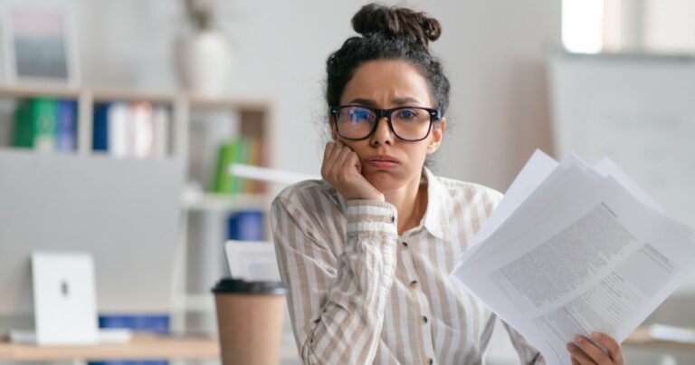 Mulher com expressão de cansaço e preocupação, usando óculos e segurando papéis em um ambiente de escritório. Ela veste uma camisa listrada e está com a mão apoiada no rosto, transmitindo sensação de sobrecarga ou estresse no trabalho. Ao fundo, um espaço de trabalho desfocado com livros e objetos de escritório