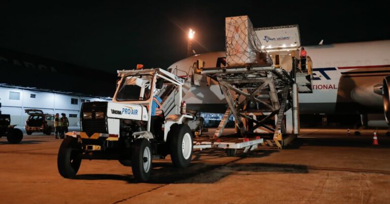 Carga do supercomputador sendo descarregada no Aeroporto do Galeão. A imagem mostra um trator de transporte de carga com a inscrição 'PRO AIR', movendo uma grande caixa de carga protegida por uma rede, com o avião de transporte visível ao fundo, em operação noturna sob iluminação artificial