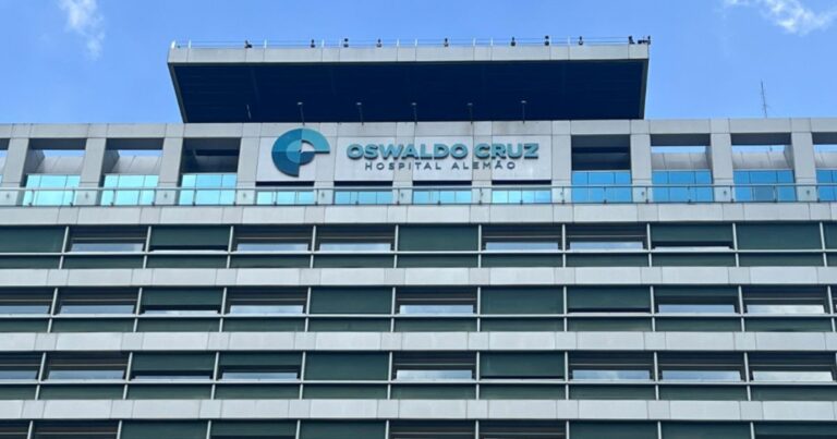 Fachada do Hospital Alemão Oswaldo Cruz, com estrutura moderna composta por linhas horizontais de janelas e um letreiro com o nome e logotipo em destaque. O céu azul ao fundo complementa a imagem, transmitindo uma atmosfera de seriedade e excelência na área da saúde (hospital, oswaldo cruz)