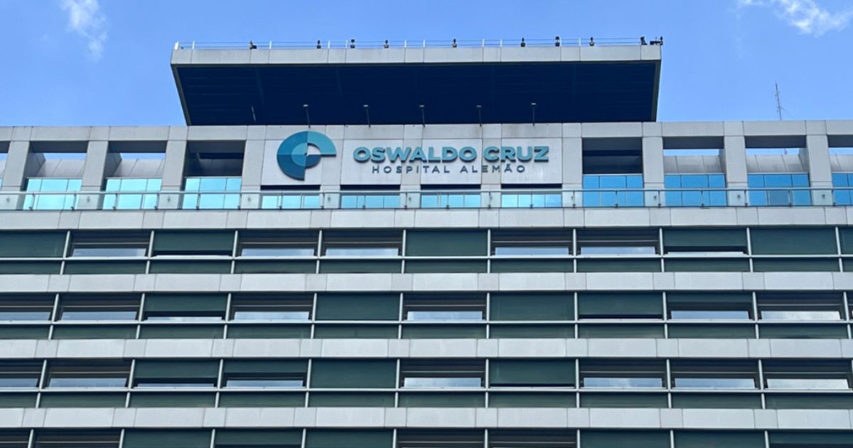 Fachada do Hospital Alemão Oswaldo Cruz, com estrutura moderna composta por linhas horizontais de janelas e um letreiro com o nome e logotipo em destaque. O céu azul ao fundo complementa a imagem, transmitindo uma atmosfera de seriedade e excelência na área da saúde (hospital, oswaldo cruz)