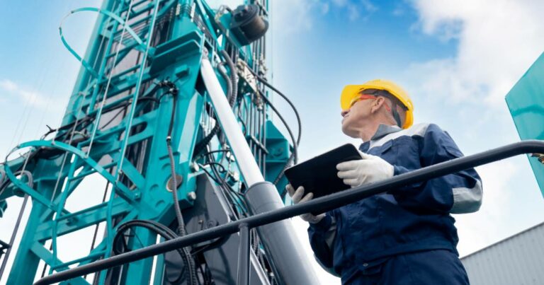 Trabalhador da indústria usando capacete amarelo e segurando um tablet enquanto inspeciona uma grande máquina industrial azul. Ele veste uniforme de proteção e luvas brancas. O céu azul ao fundo complementa o ambiente industrial ao ar livre, política, indústria