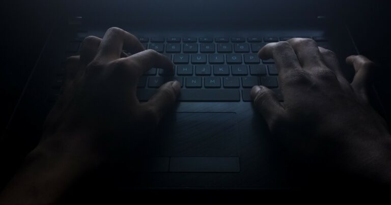 Mãos digitando em um teclado de laptop em um ambiente escuro, iluminadas por uma luz fraca que destaca o cenário misterioso e sombrio. A imagem evoca o conceito do lado obscuro da tecnologia, como cibercrimes ou atividades clandestinas, valor