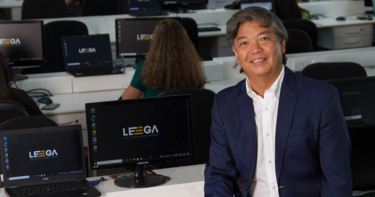 Newton Ide, executivo da Leega, posando em um ambiente corporativo com computadores ao fundo, todos exibindo o logotipo da empresa Leega nas telas. Ele está sorrindo, vestindo um blazer azul e camisa branca, transmitindo profissionalismo e liderança (newton ide, leega))