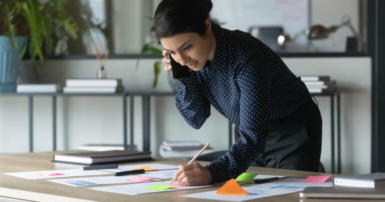 Profissional em um ambiente de escritório analisando documentos e gráficos sobre uma mesa, enquanto fala ao telefone. Ela usa uma camisa escura de bolinhas e segura um lápis, anotando em folhas com post-its coloridos espalhados, sugerindo um planejamento estratégico ou organização de tarefas. Ao fundo, o escritório apresenta prateleiras com livros e plantas, criando um ambiente de trabalho focado e organizado
