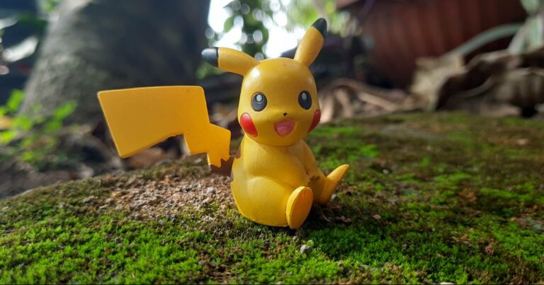 Figura de um Pikachu, pokémon, em miniatura, sentado sobre musgo em um ambiente natural, com um fundo desfocado de plantas e árvores.