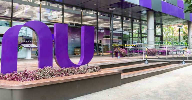 Entrada moderna do escritório do Nubank, com um grande letreiro roxo em forma das letras 'nu' em destaque. A fachada de vidro reflete a arquitetura contemporânea e o ambiente externo, com áreas de convivência e vegetação ao redor, criando um espaço acolhedor e inovador