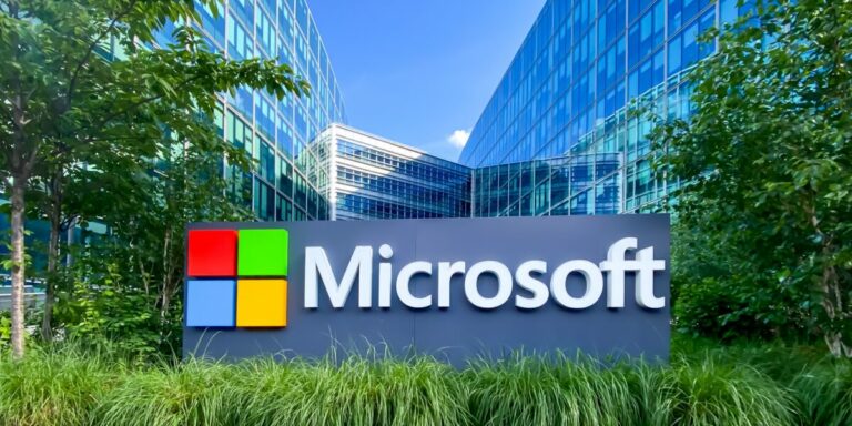 Logotipo da Microsoft em destaque em um painel em frente a um moderno complexo de escritórios com fachada de vidro. O ambiente é cercado por vegetação e árvores, com céu azul ao fundo, criando uma atmosfera corporativa e sustentável, cursos