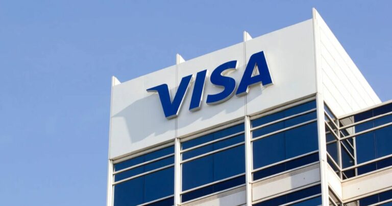 Fachada de um prédio corporativo da Visa com o logotipo azul da empresa em destaque sobre um fundo branco, contra um céu azul claro. A estrutura moderna do edifício é composta por janelas de vidro refletivo (visa, vai de visa)