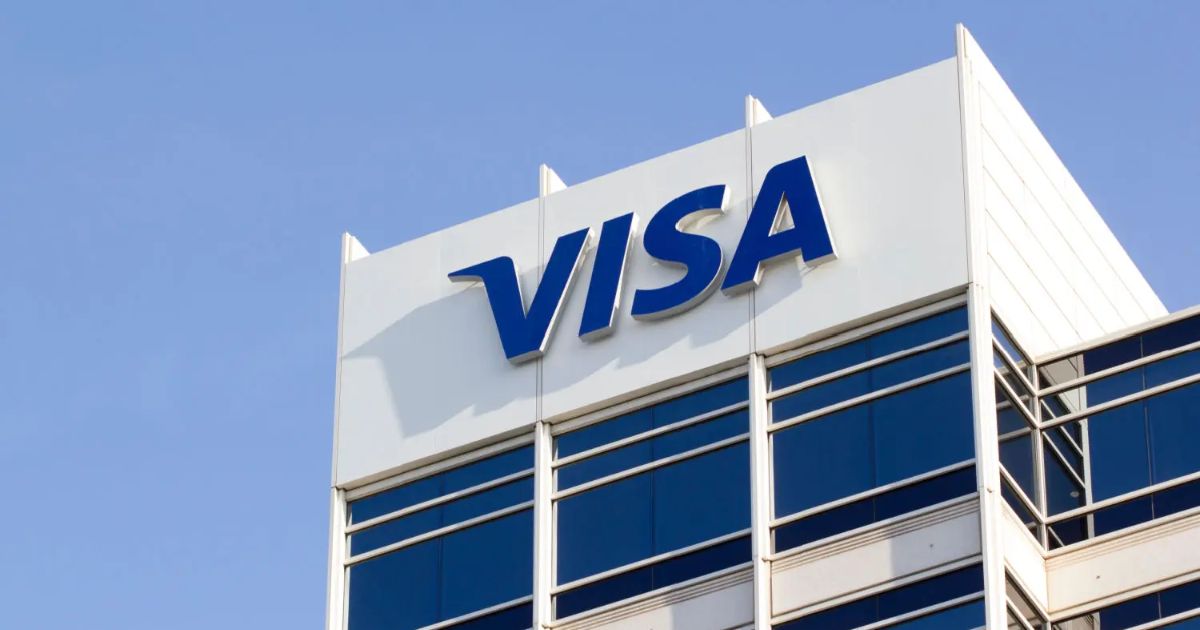 Fachada de um prédio corporativo da Visa com o logotipo azul da empresa em destaque sobre um fundo branco, contra um céu azul claro. A estrutura moderna do edifício é composta por janelas de vidro refletivo (visa, vai de visa)