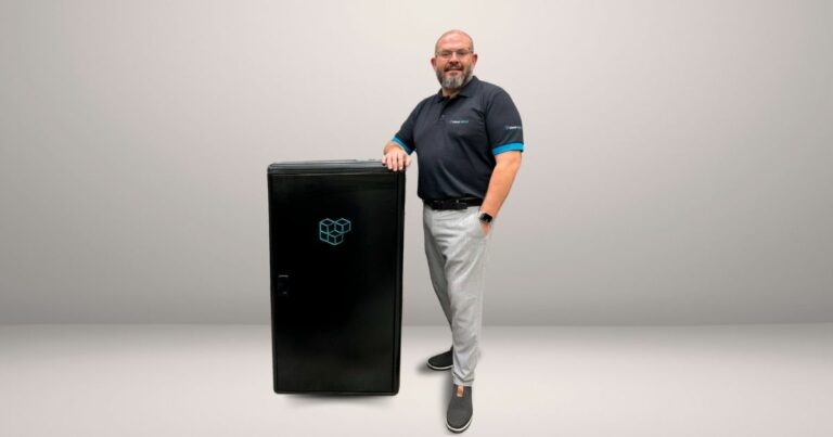 A imagem apresenta André Machado, head de pesquisa, desenvolvimento e inovação da cloudvBOX, posando ao lado do vBOX EDGE, a solução desenvolvida pela empresa. Ele está vestido com uma camisa polo preta, com detalhes em azul nas mangas e o logotipo da empresa, além de calça clara e sapatos casuais escuros. O vBOX EDGE, exibido ao seu lado, é um equipamento de cor preta com o logotipo da cloudvBOX em destaque, ilustrando o design compacto e funcional do micro data center. O fundo é neutro, direcionando toda a atenção para André e o equipamento.