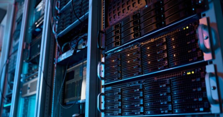 A imagem mostra um ambiente de data center com diversos servidores montados em racks. Os equipamentos estão organizados em fileiras verticais, com luzes indicadoras acesas em alguns dos dispositivos, sugerindo que estão em operação. A iluminação é predominantemente azulada, com tons sutis de roxo, criando uma atmosfera tecnológica e moderna. Cabos e componentes conectam os servidores, refletindo a infraestrutura robusta necessária para armazenamento e processamento de dados em larga escala. Esse cenário é típico de locais utilizados para operações de TI críticas, como hospedagem de sites, análise de dados, inteligência artificial e outras aplicações de computação avançada. (emissões, brasil)