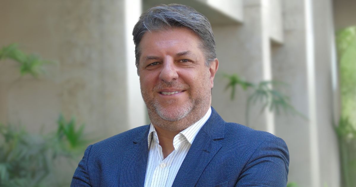 Dennis Milan, diretor de tecnologia, inovação e digital da Tokio Marine, sorrindo em um ambiente externo com plantas e paredes claras ao fundo. Ele está vestido com um blazer azul e camisa branca, transmitindo profissionalismo e confiança