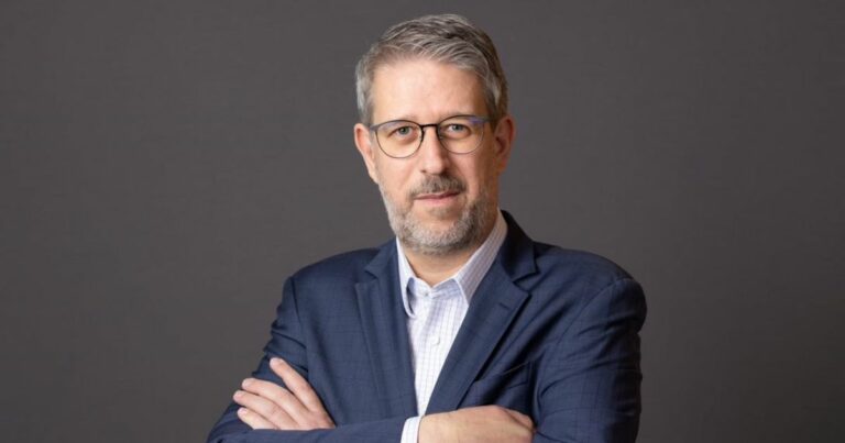 Na foto, Juliano de Conti aparece posicionado diante de um fundo neutro, vestindo um paletó azul-escuro sobre uma camisa social clara. Ele mantém os braços cruzados e exibe uma expressão séria e confiante, típica de um executivo seguro de suas decisões. Seu olhar direcionado à câmera transmite profissionalismo e compostura, reforçando a imagem de liderança e competência.