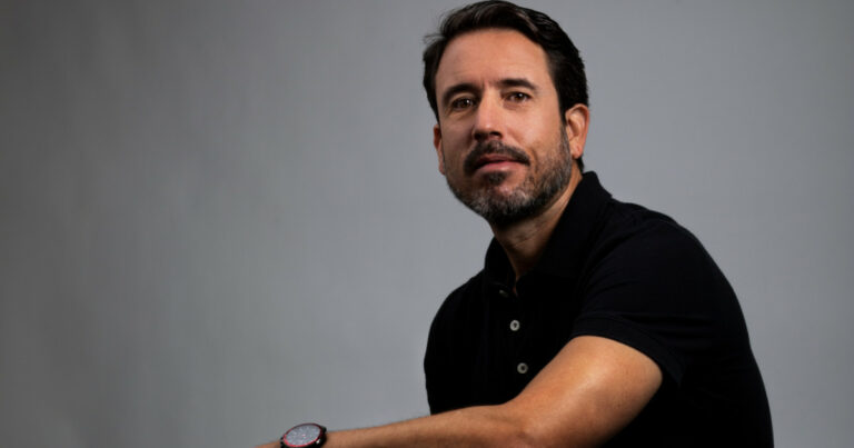 Marcelo Torres, cofundador e COO da Kakau Seguros, posando com expressão confiante em um fundo cinza. Ele veste uma camisa preta e um relógio vermelho, transmitindo modernidade e profissionalismo