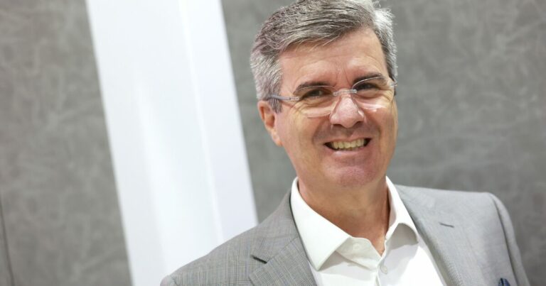 A imagem mostra um homem de óculos e cabelo grisalho, sorrindo para a câmara. Ele veste um fato cinzento claro e uma camisa branca, transmitindo uma aparência profissional e amigável. O fundo é neutro, com tons de cinza e branco, destacando o sujeito na fotografia. A iluminação é suave, criando um ambiente limpo e corporativo.