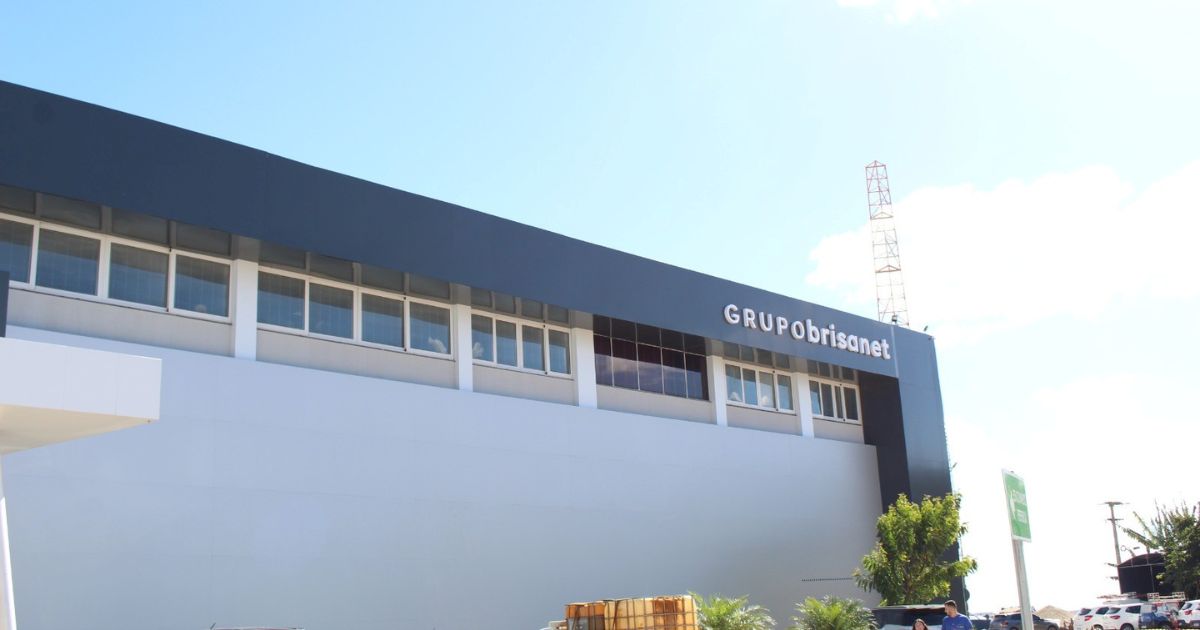Prédio comercial moderno com fachada branca e detalhes em cinza-escuro. O edifício apresenta várias janelas retangulares alinhadas na parte superior. Há uma placa com o nome 'GRUPO brisanet' em destaque na lateral do prédio. No fundo, um céu azul com poucas nuvens e uma torre de telecomunicação visível. Na parte inferior direita, há árvores e algumas estruturas menores, sugerindo um ambiente urbano.