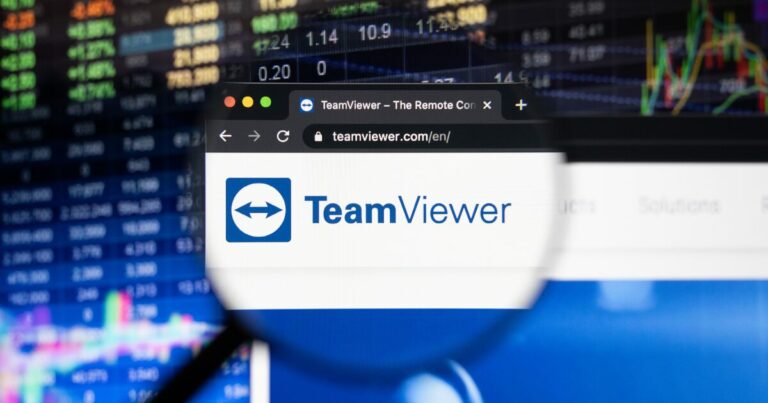 Imagem da interface do navegador de internet mostrando a página inicial do site da TeamViewer (teamviewer.com/en/), destacada por uma lupa sobre o logotipo da empresa, composto por um ícone azul com setas brancas e o texto "TeamViewer". Ao fundo, gráficos financeiros e números desfocados sugerem um contexto de negócios e tecnologia.
