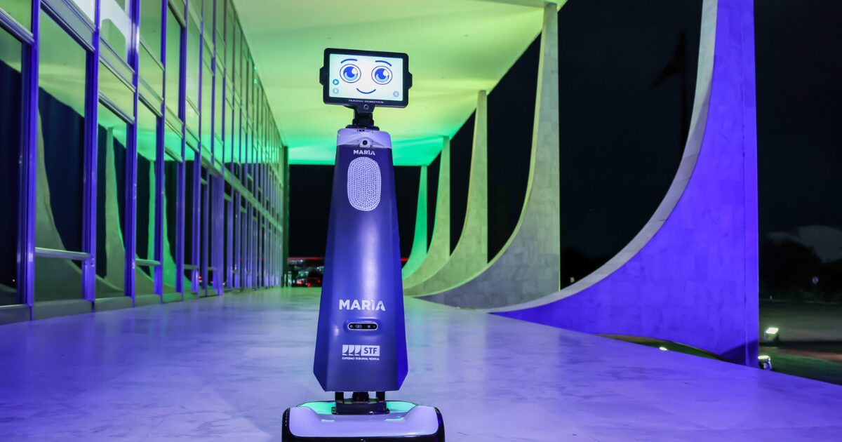 Um robô chamado "Maria", posicionado em um corredor futurista iluminado por luzes verdes e roxas. O robô tem um design moderno e cilíndrico, com um rosto digital sorridente exibido em uma tela no topo. Na frente do corpo do robô, aparecem as palavras “MARIA” e o logotipo do “STF”, indicando que ele pertence ao Supremo Tribunal Federal. O ambiente tem grandes colunas de concreto com linhas curvas características, indicando que a imagem foi tirada no prédio do STF em Brasília, Brasil.