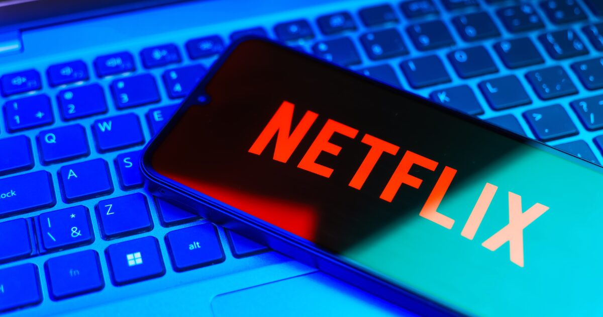 Um smartphone exibindo o logotipo da Netflix em letras vermelhas está apoiado sobre um teclado de computador iluminado com tons de azul.