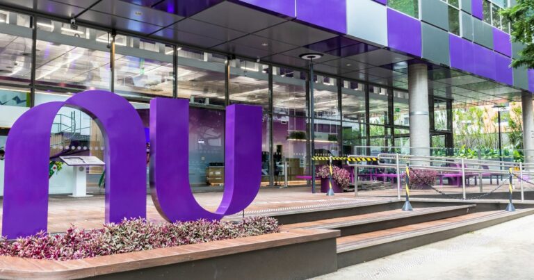 A imagem mostra a fachada de um edifício moderno, com amplas janelas de vidro e painéis externos na cor roxa e cinza. Em destaque, há uma grande escultura roxa em formato de "n" e "u" estilizados, que parece representar o logo do Nubank. Em frente à entrada do edifício, há uma rampa de acesso com corrimões metálicos e faixas amarelas e pretas de segurança. Vaso com plantas roxas e verdes decoram o espaço, harmonizando com o design moderno e colorido do local. Ao fundo, pode-se ver mais vegetação e uma área externa com bancos e mesas. (OXXO)