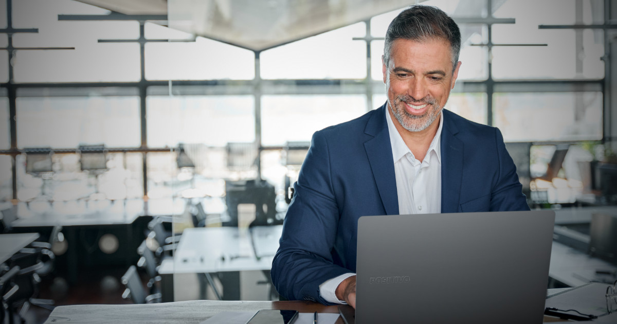A imagem mostra um homem sorrindo enquanto trabalha em um laptop em um ambiente de escritório moderno e iluminado. Ele está usando um terno azul escuro e uma camisa branca. O fundo do escritório apresenta janelas grandes, proporcionando luz natural ao espaço, e mesas de trabalho vazias. O laptop que ele está utilizando tem a marca "Positivo" visível em sua parte superior.