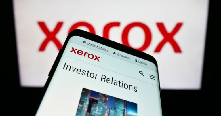 A imagem mostra um smartphone exibindo a página "Investor Relations" no site da Xerox. No fundo, aparece o logotipo da Xerox em vermelho, ampliado e desfocado. O site visível no celular inclui opções de navegação como "United States," "Account," e "Log In," além de uma barra de busca. A composição destaca o foco em informações corporativas e a marca Xerox.