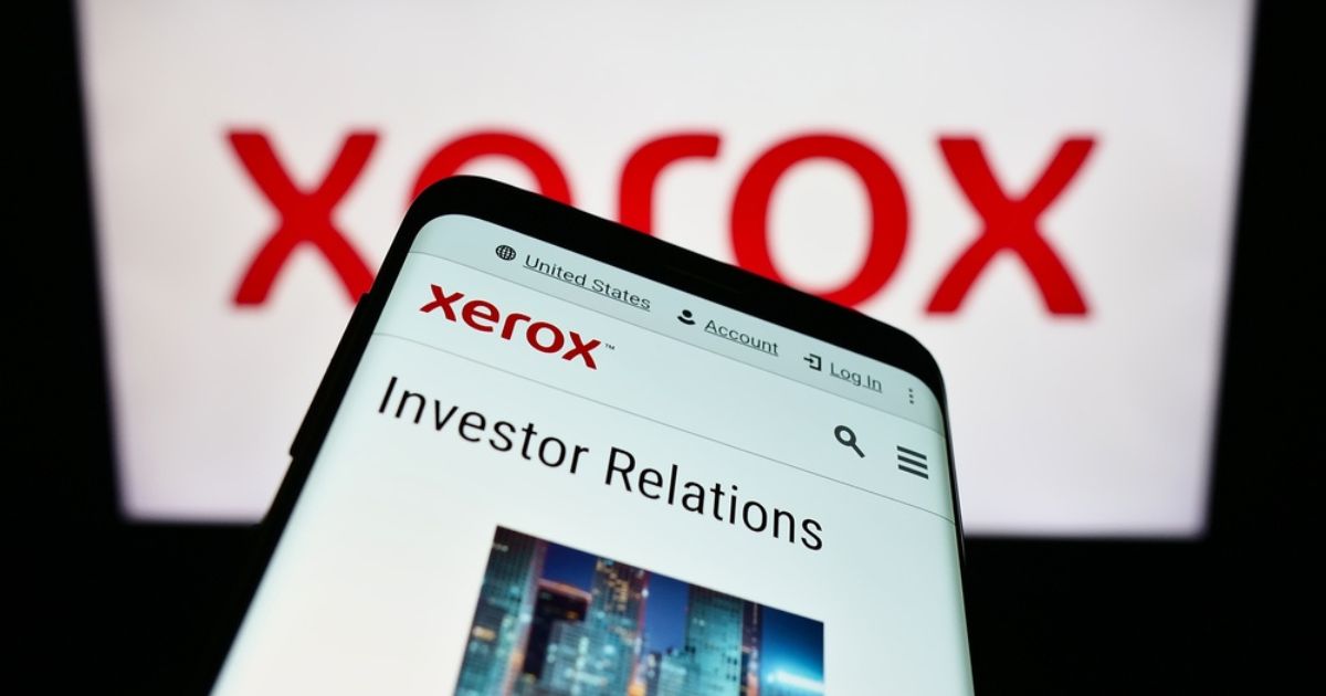 A imagem mostra um smartphone exibindo a página "Investor Relations" no site da Xerox. No fundo, aparece o logotipo da Xerox em vermelho, ampliado e desfocado. O site visível no celular inclui opções de navegação como "United States," "Account," e "Log In," além de uma barra de busca. A composição destaca o foco em informações corporativas e a marca Xerox.