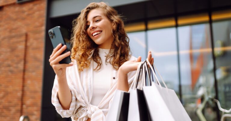 A imagem mostra uma mulher sorrindo enquanto segura várias sacolas de compras em uma das mãos e um smartphone na outra. Ela está ao ar livre, em frente a um estabelecimento comercial moderno, com janelas grandes ao fundo. A mulher tem cabelos cacheados e ruivos, e veste uma blusa branca com listras. Ela parece feliz e satisfeita com suas compras, sugerindo uma experiência de compra positiva, possivelmente em um ambiente de varejo ou retailtech (compras, varejo, celular, sacolas, mulher, ruiva, smartphone, retailtechs)