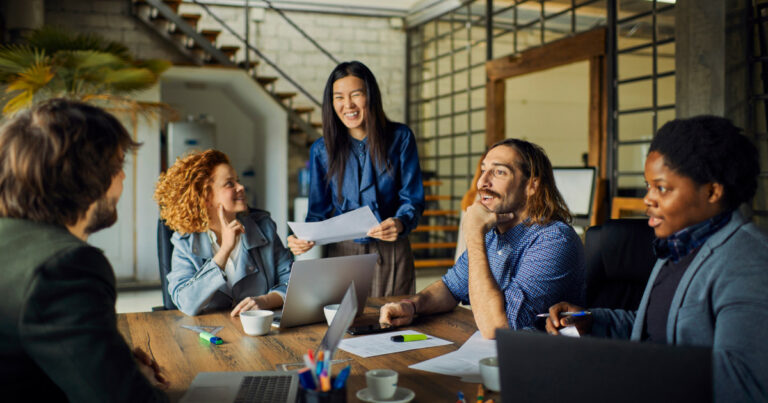 A imagem mostra um grupo diverso de profissionais em uma reunião colaborativa. Cinco pessoas estão sentadas ao redor de uma mesa, sorrindo e interagindo. Uma mulher está em pé, segurando alguns papéis, enquanto os outros estão ouvindo atentamente e sorrindo. Há duas mulheres e três homens, representando uma diversidade de etnias e estilos. A mesa está organizada com laptops, canetas, e papéis, criando um ambiente descontraído e produtivo. O ambiente é moderno, com iluminação natural e uma parede de tijolos ao fundo, sugerindo um espaço criativo de trabalho (diversidade, ento)