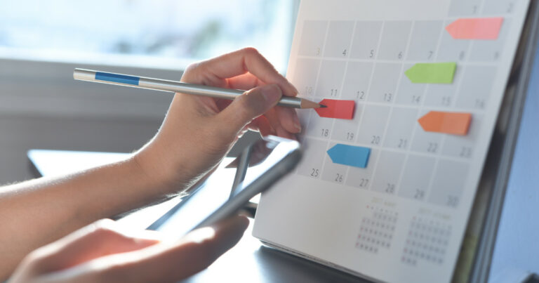 Pessoa utilizando um calendário de mesa para planejar datas importantes, marcando um dia com uma etiqueta colorida enquanto segura um celular. A imagem destaca organização e gerenciamento de tempo em um ambiente iluminado por luz natural, trabalho