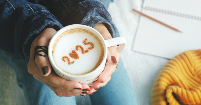Mãos segurando uma xícara de café com espuma, onde está escrito "2025" em canela. Ao fundo, aparecem um caderno com um lápis e um tecido amarelo, compondo um ambiente acolhedor e tranquilo, simbolizando a chegada do Ano Novo de 2025. As mãos têm unhas pintadas e um anel, sugerindo um momento de reflexão ou celebração